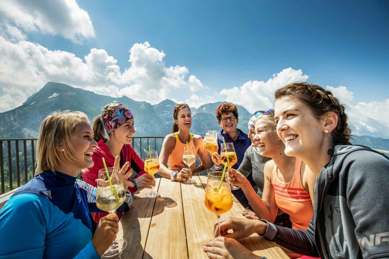 Sieben Frauen trinken lachend Aperol auf der Sonnenterasse der Jenneralm