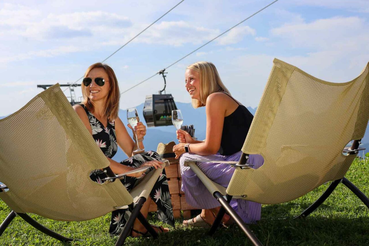 Zwei Frauen blicken im Liegestuhl sitzend und lächeln in eine Richtung und haben dabei ein Glas Prosecco in der Hand